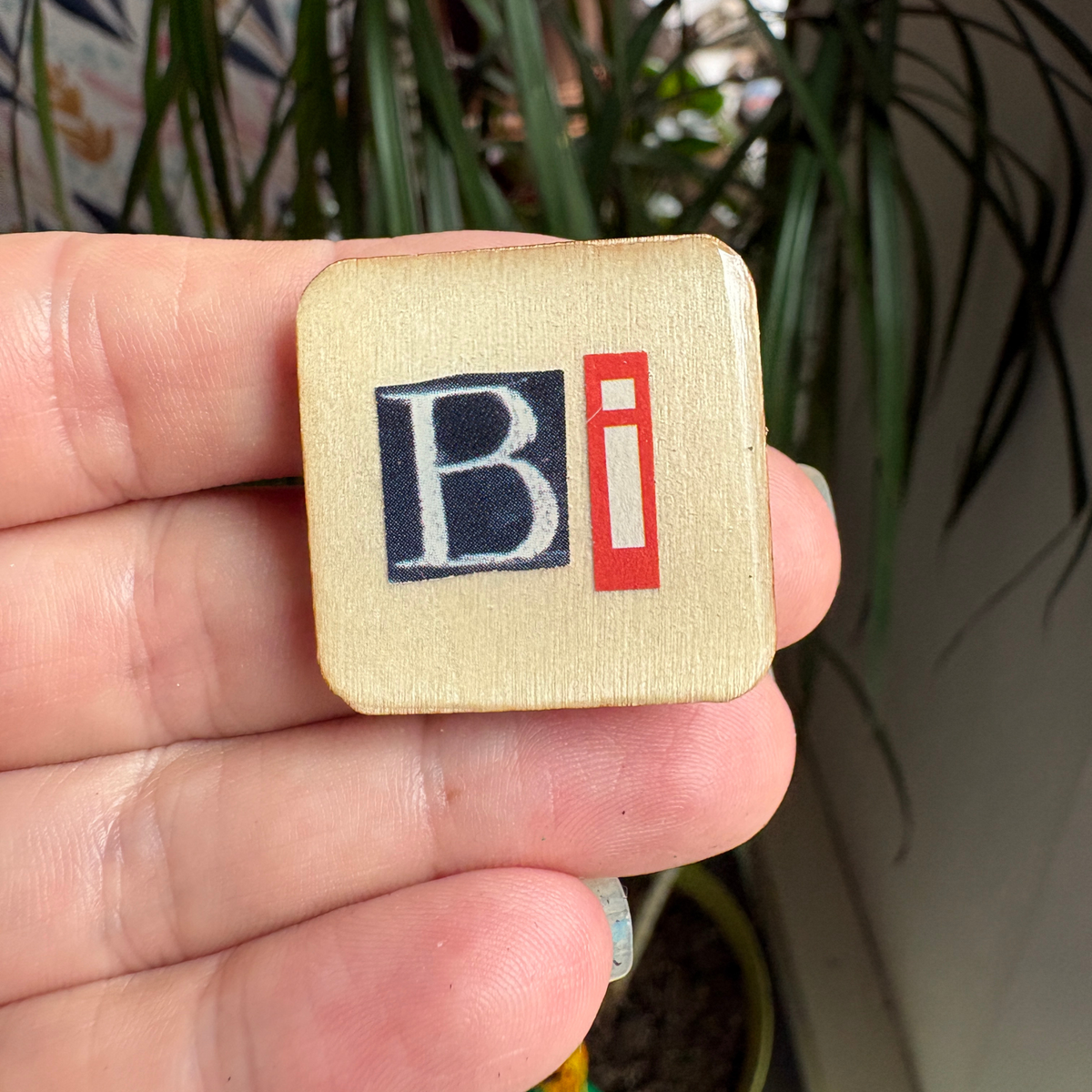 Hand holding a wooden pin with 'Bi' letters, blurred plant in the background