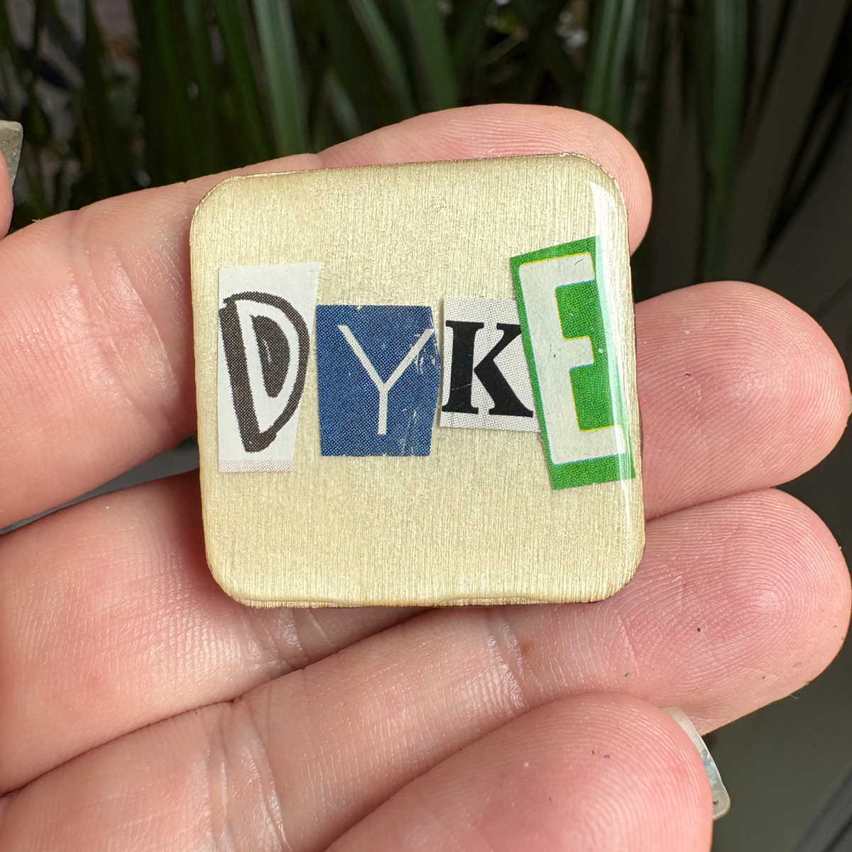 Small square pin with cut-out letters spelling 'DYKE' held in a hand.