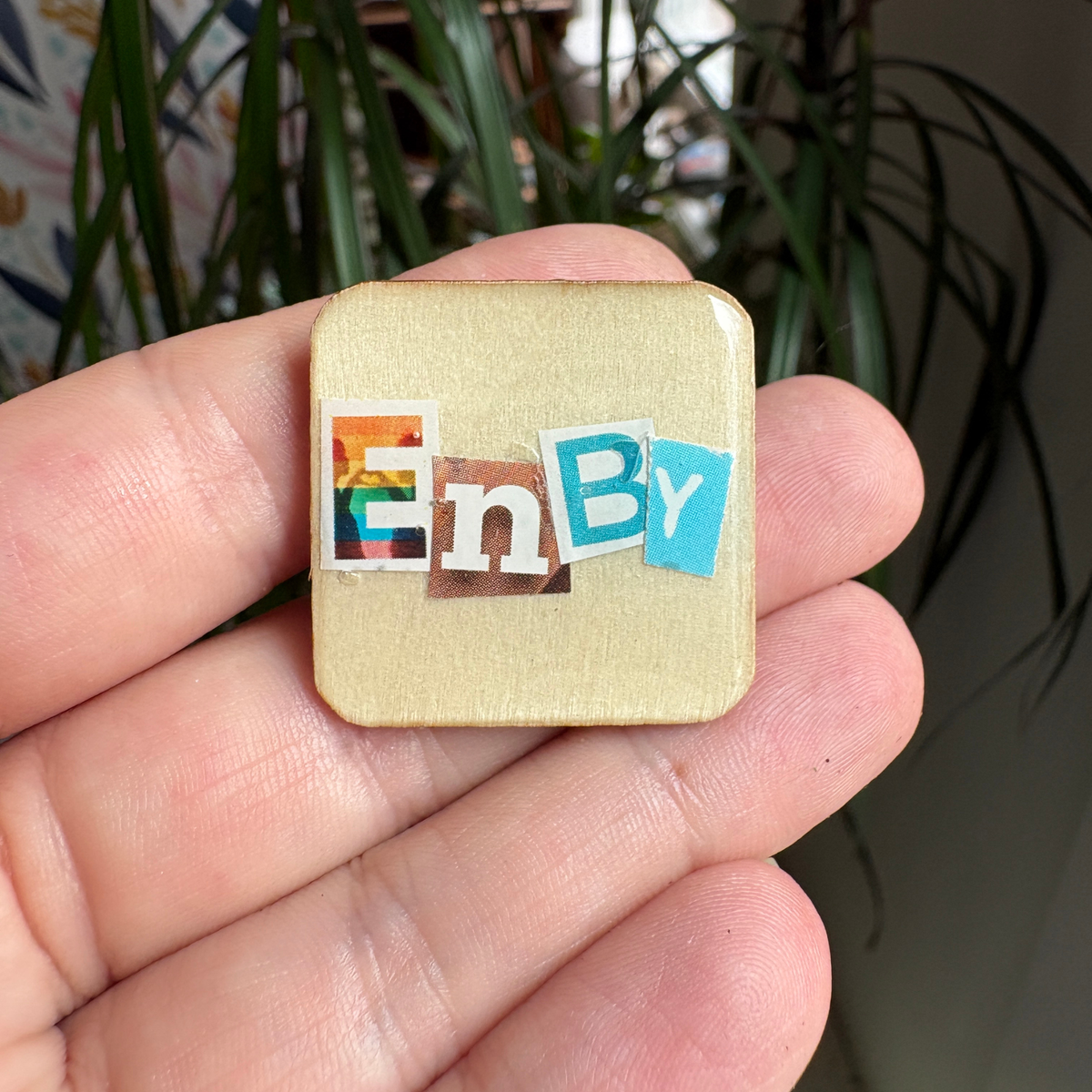 Small square pin with colorful letters 'EnBy' held in a hand against a plant background
