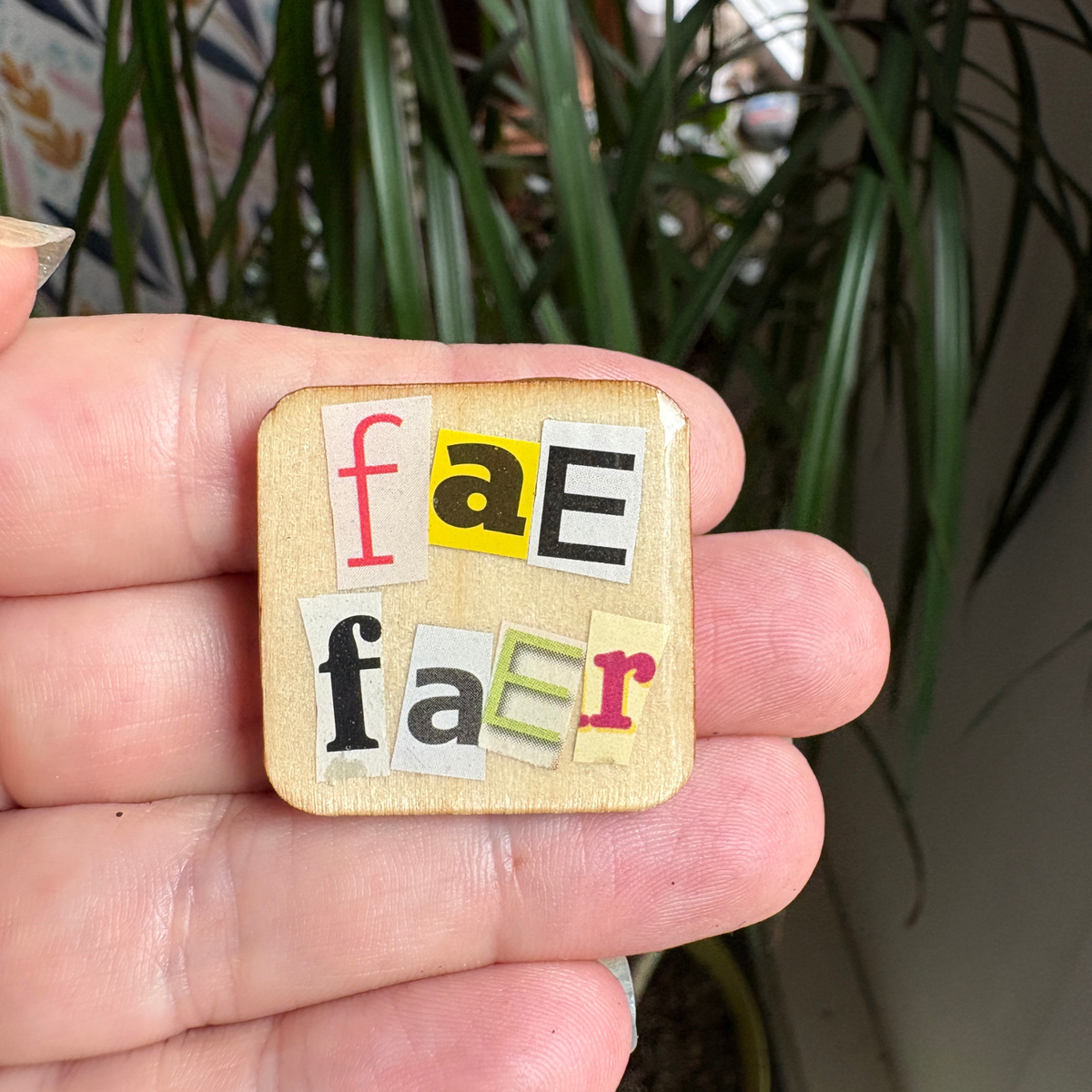 Wooden square pin with colorful letters spelling 'fae' and 'faer' held in a hand with a plant in the background