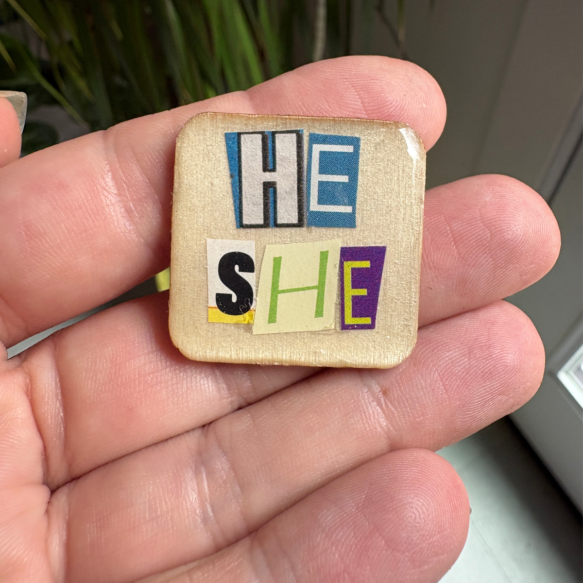 Small square wooden pin with 'HE SHE' in colorful letters held in a hand.