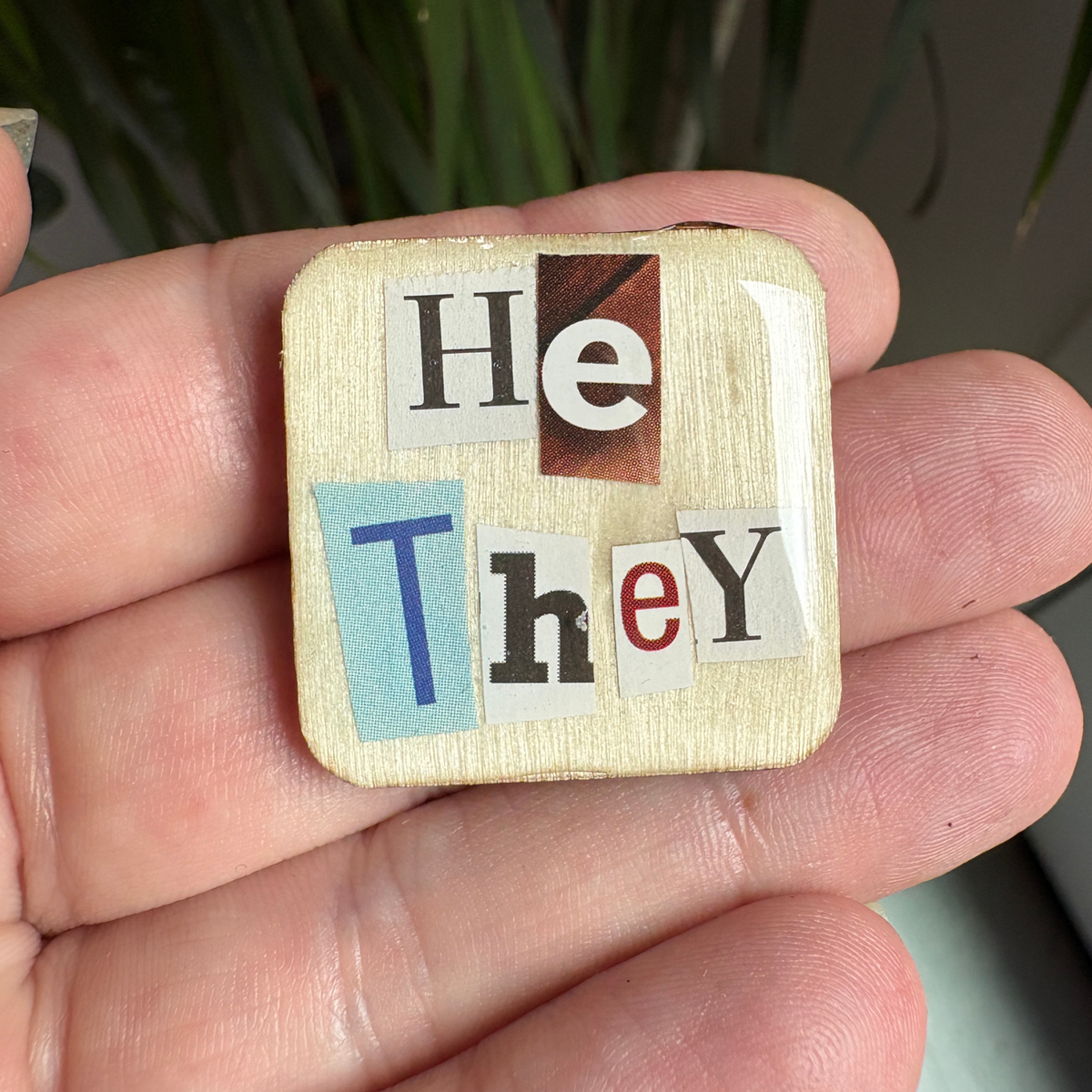 Wooden square pin with letters forming 'He They' held in a hand
