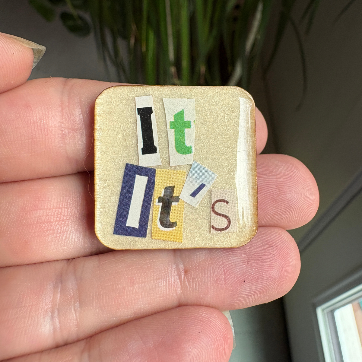 Square wooden pin with cut-out letters spelling 'It it's' held in a hand.