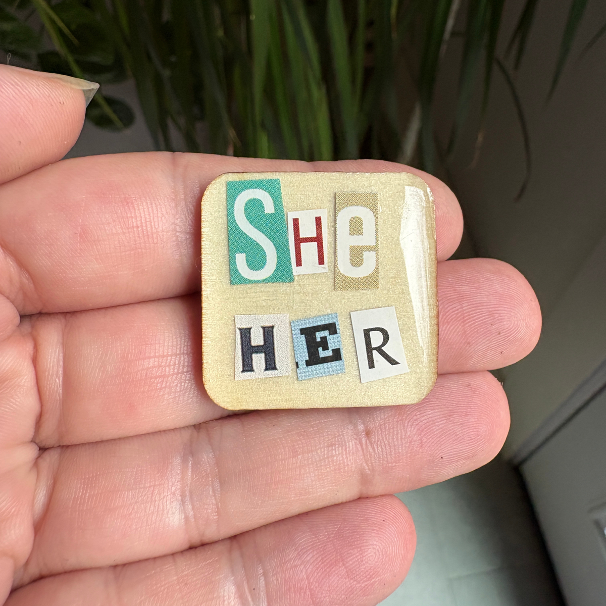 Small square pin with 'SHE/her' text held in a hand against a blurred background