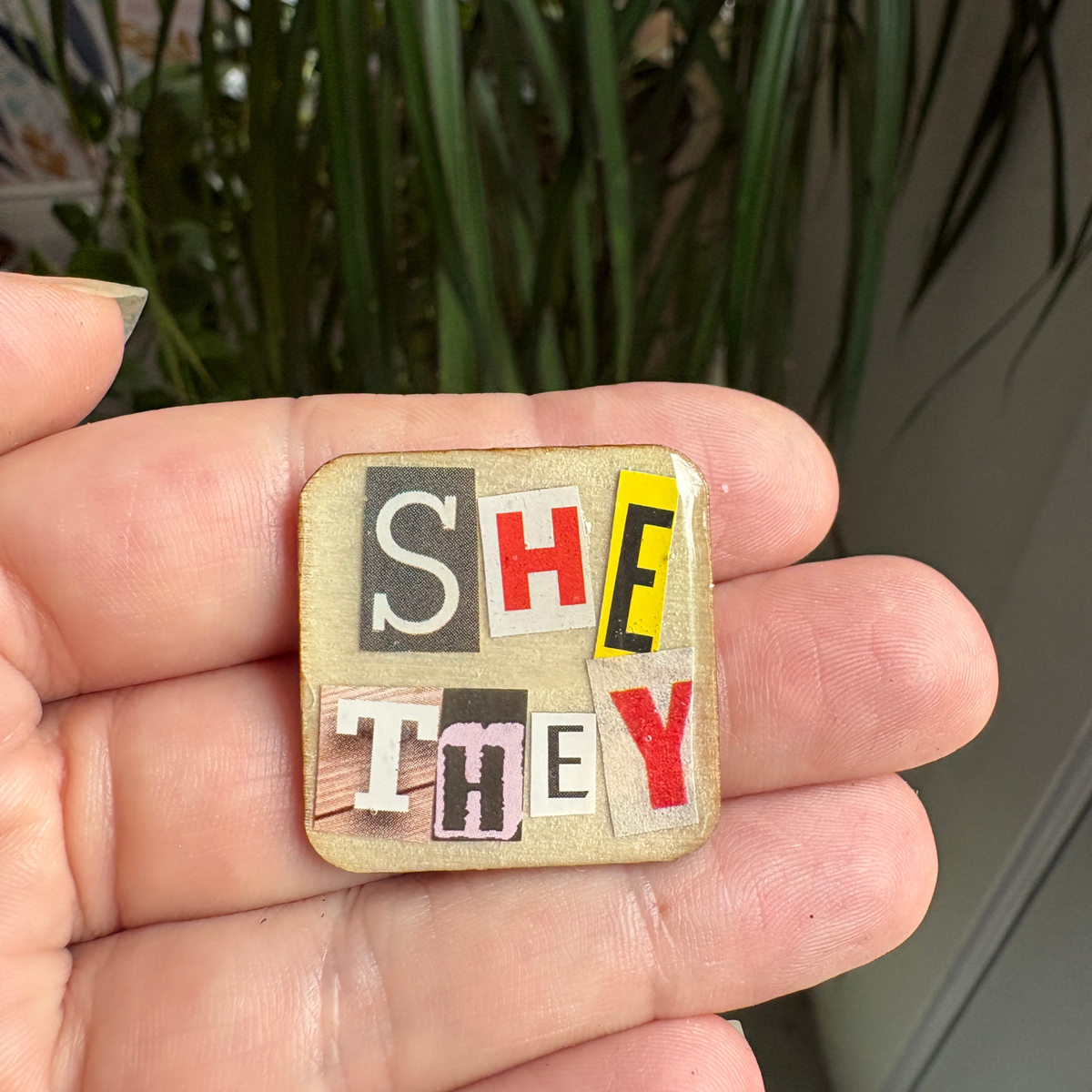 Small square patch with 'SHE' and 'THEY' letters held in a hand, with a plant in the background.