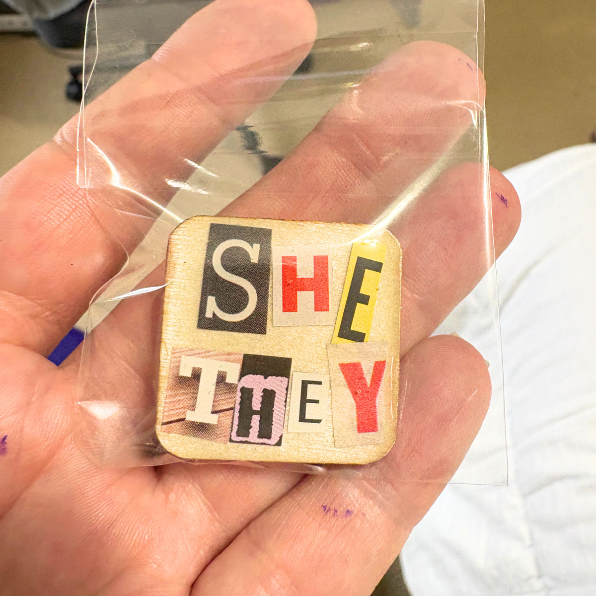 Wooden square pin with letters spelling 'SHE' and 'THEY' held in a hand.