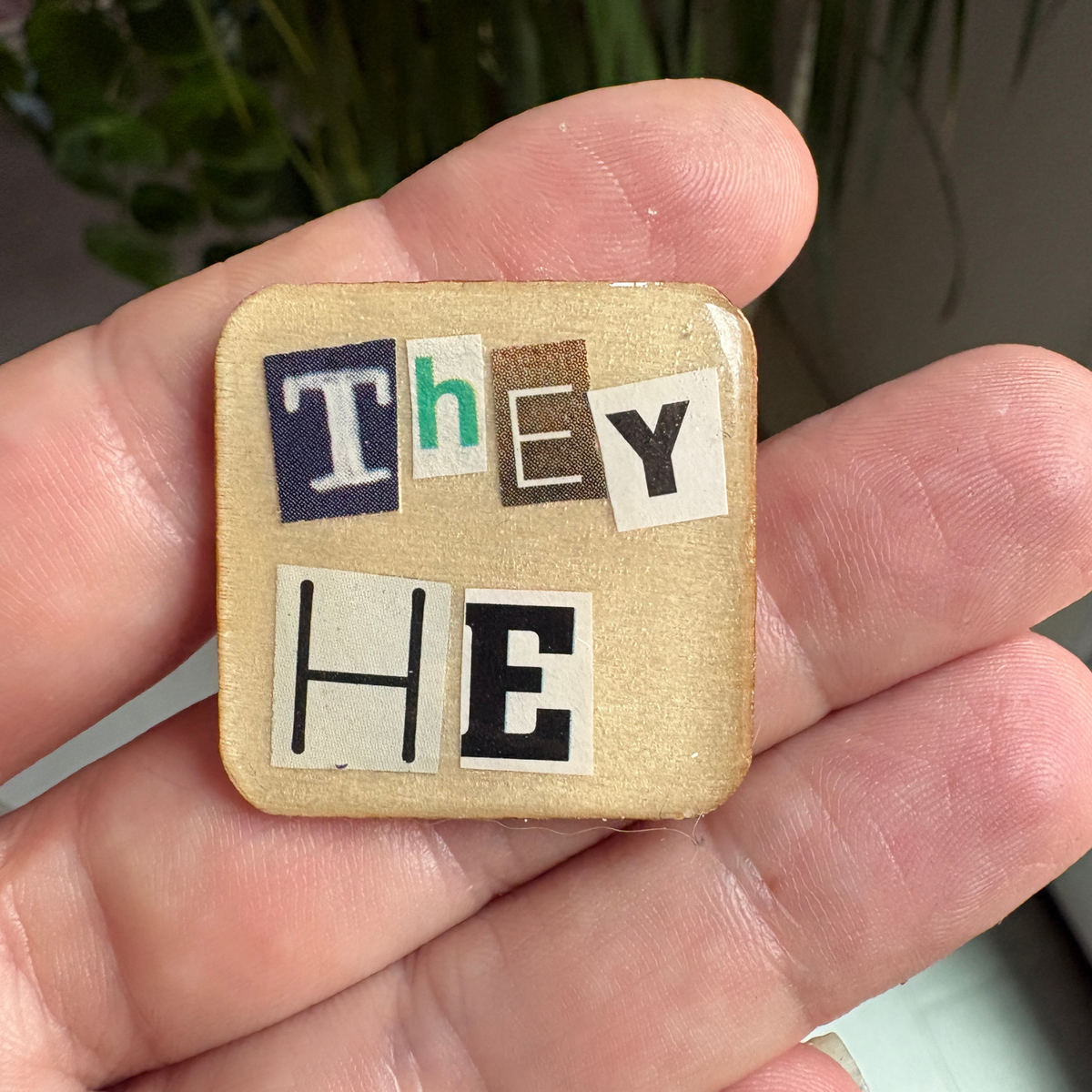 Small wooden square pin with letters spelling 'THEY HE' held in a hand.