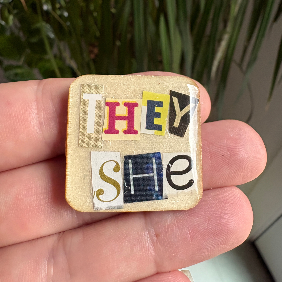 Small square pin with 'THEY SHE' text held in a hand against a blurred plant background