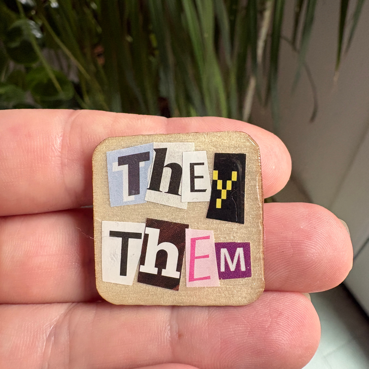 Small square pin with 'They' and 'Them' text held between fingers against a blurred plant background