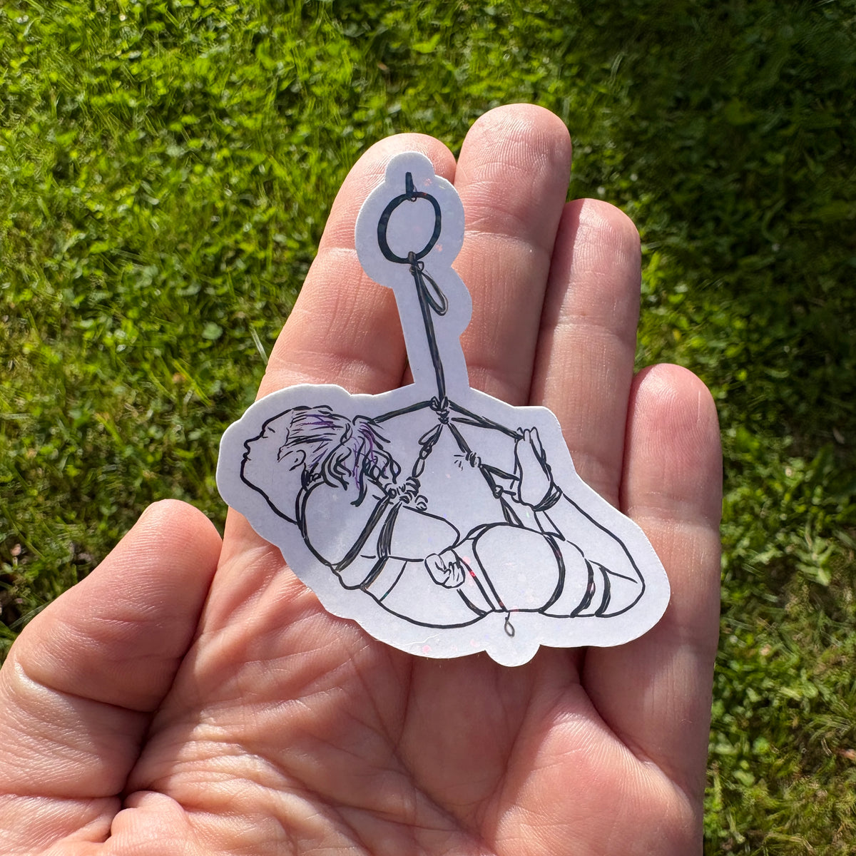 Hand holding a black and white sticker of a person in a hogtie rope suspension against a grassy background