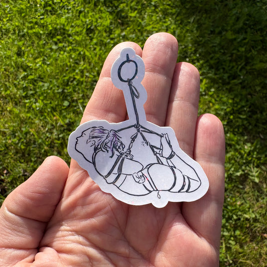 Hand holding a black and white sticker of a person in a hogtie rope suspension against a grassy background