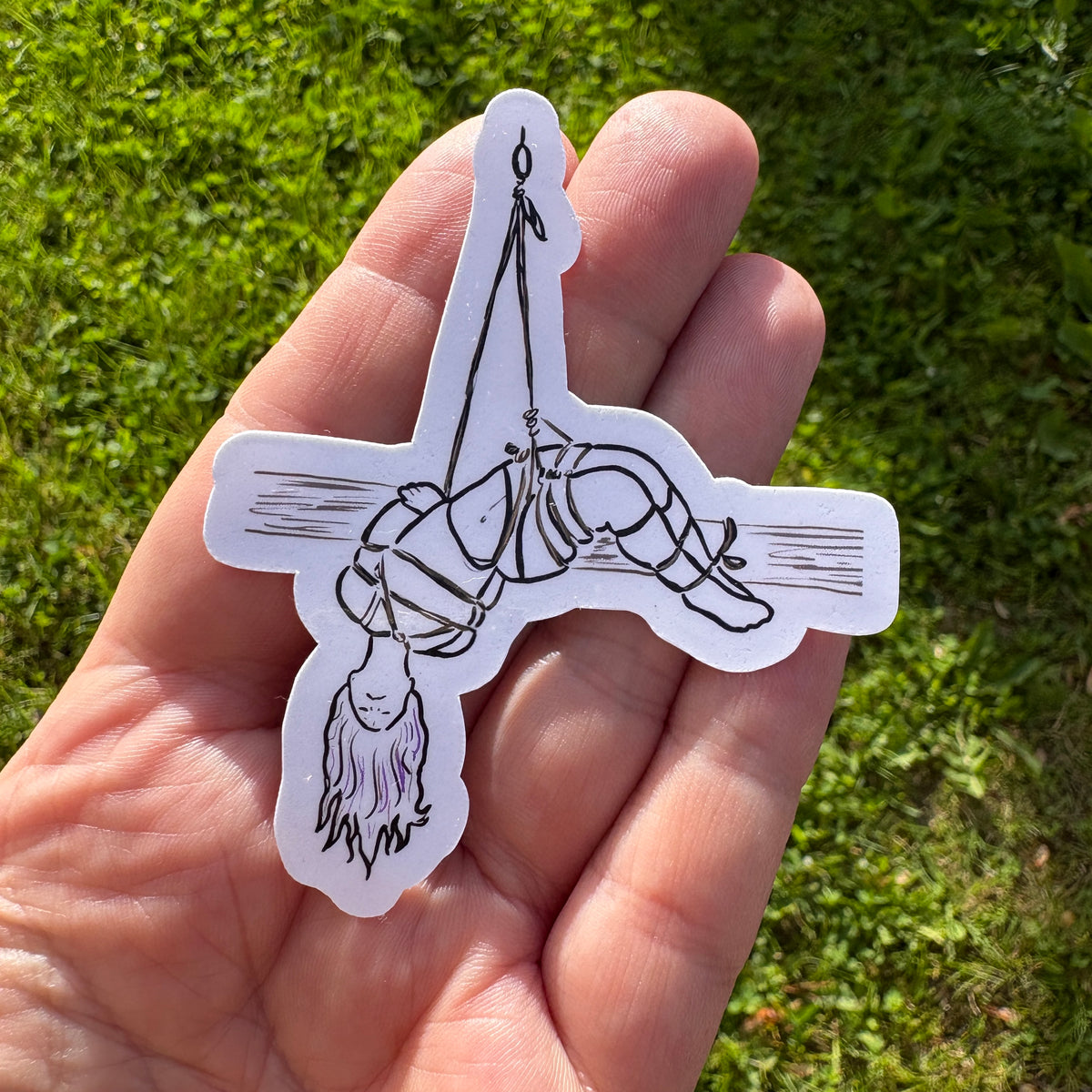 Hand holding a sticker of a person hanging from a rope harness against a grassy background