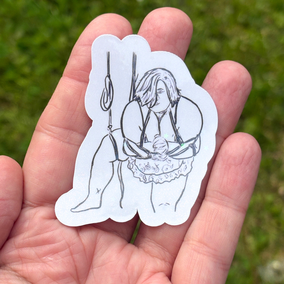 Hand holding a white sticker with a black line drawing of a person in a rope suspension against a green grass background.