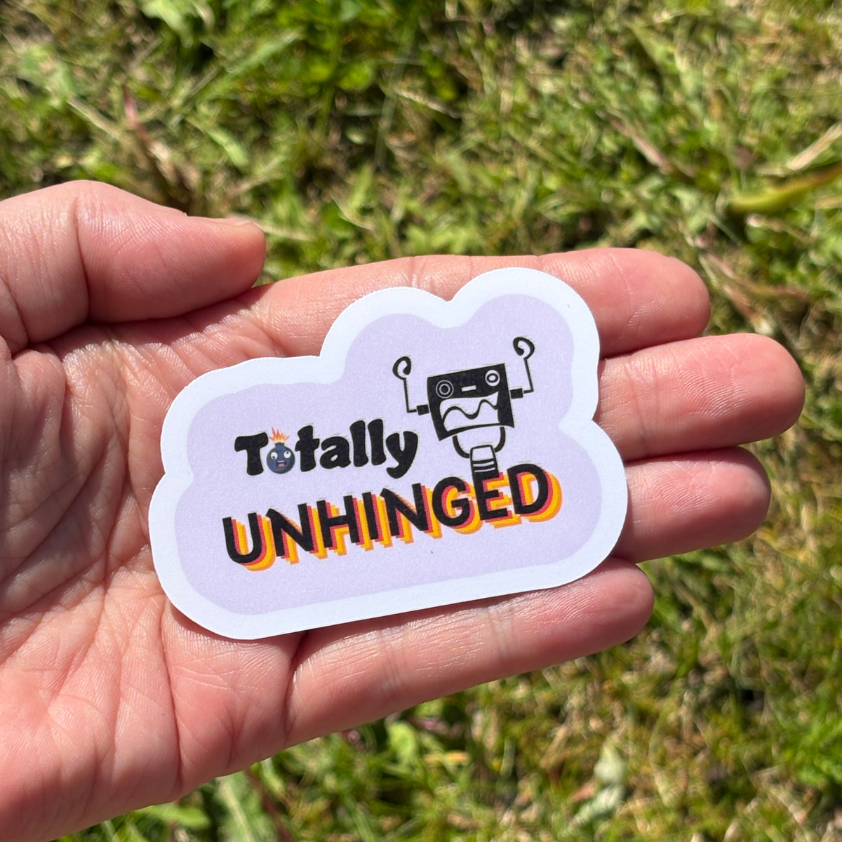 Hand holding a 'Totally UNHINGED' sticker with a robot graphic against a grassy background