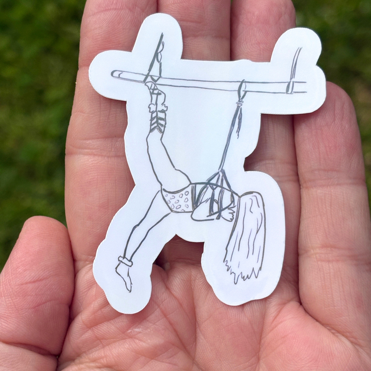 Hand holding a white sticker with a black line drawing of a person in a rope suspension against a blurred green background.