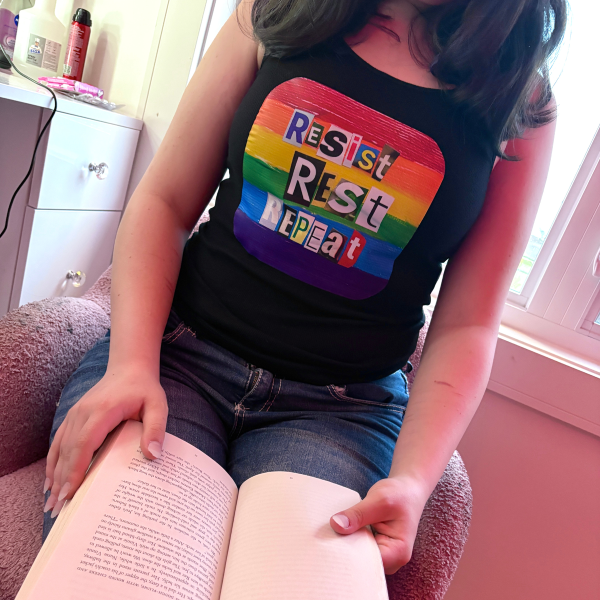 Person wearing a black tank top with rainbow-colored box containing the statement Resist Rest Repeat in collage lettering. The person is sitting on a couch holding an open book.