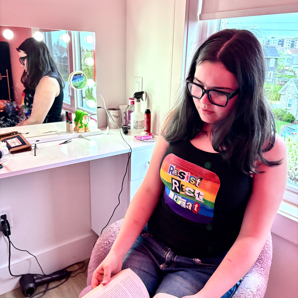 Person wearing a black tank top with rainbow-colored box containing the statement Resist Rest Repeat in collage lettering. The person is sitting on a chair holding an open book.