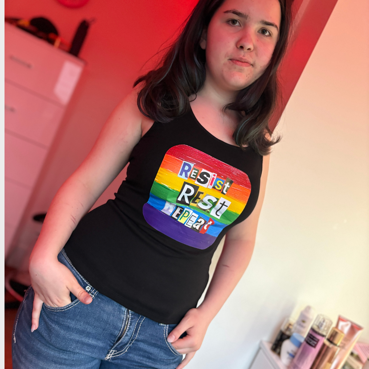 Person wearing a black tank top with rainbow-colored box containing the statement Resist Rest Repeat in collage lettering. 