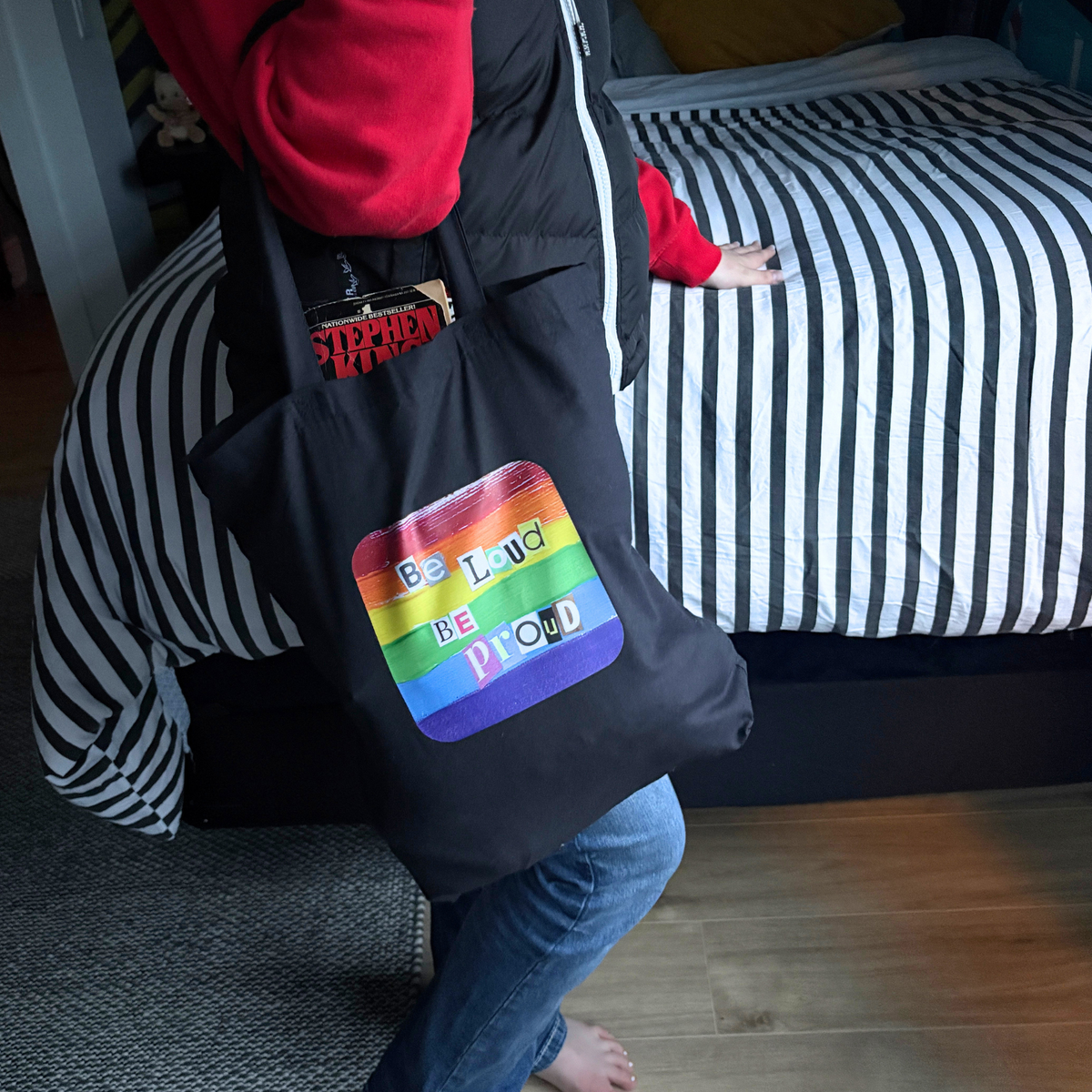 Black tote bag with rainbow pride design and text 'Be Loud Be Proud' held by a person.