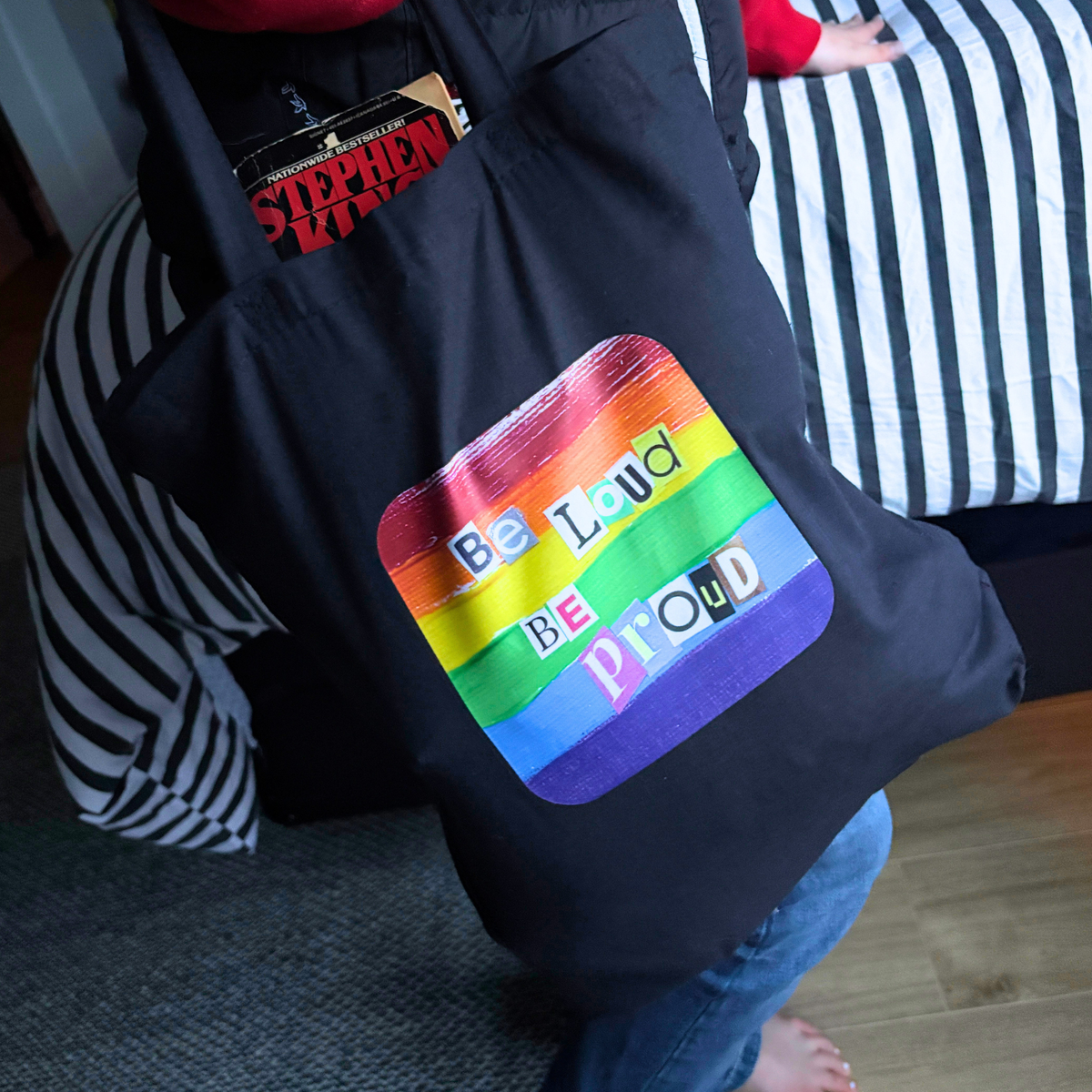 Black tote bag with rainbow pride design and text 'Be Loud Be Proud' held by a person.