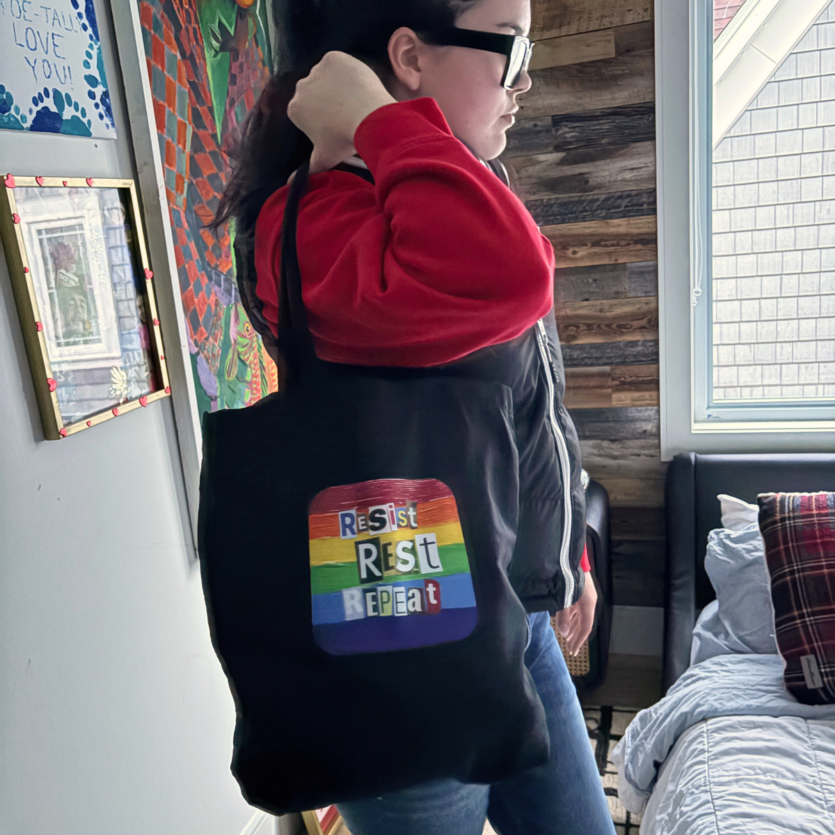 Person holding a black tote with a rainbow 'Resist Rest Repeat ' logo
