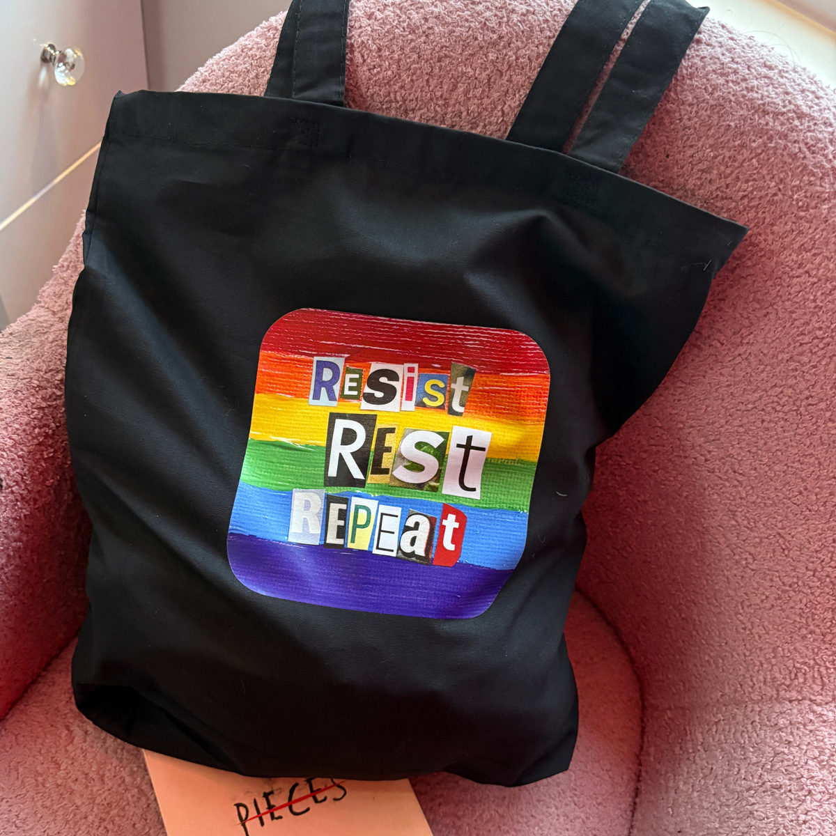 Black tote bag with a rainbow-colored patch featuring the text 'Resist Rest Repeat' on a pink chair.