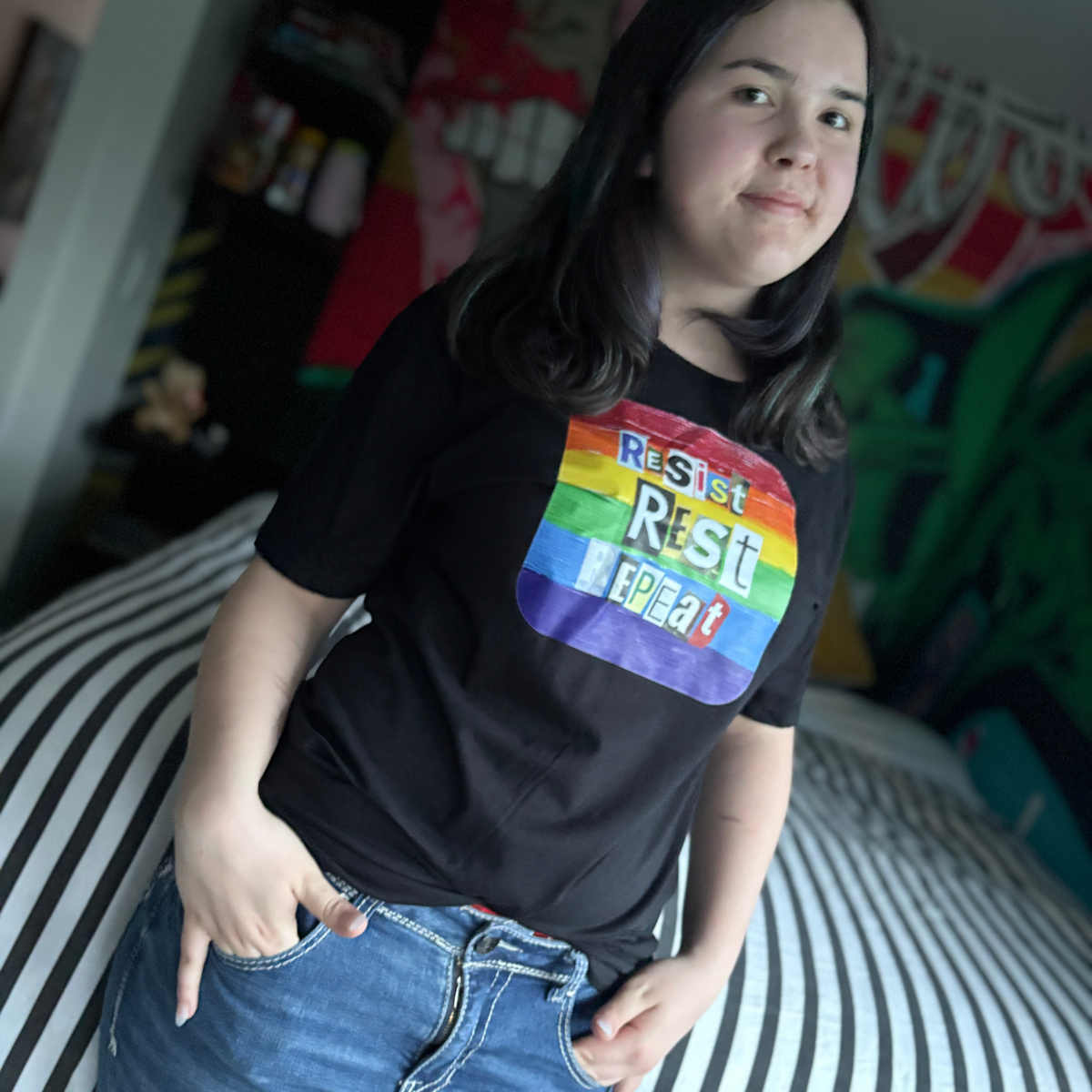 Person wearing a black t-shirt that reads Resist Rest Repeat in collage lettering on a rainbow background.