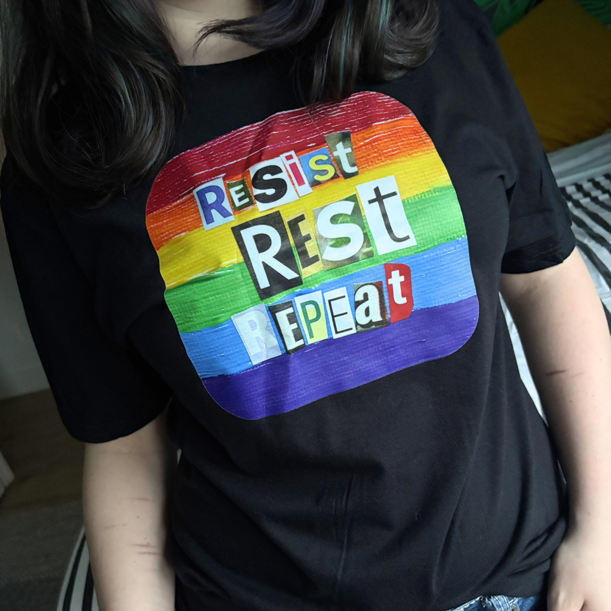 Person wearing a black t-shirt that reads Resist Rest Repeat in collage lettering on a rainbow background